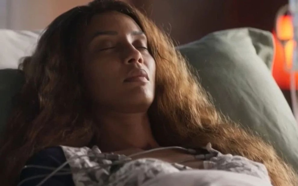 Taís Araujo, caracterizada como Clarice, está deitada em um leito hospitalar, com os olhos fechados e o semblante sereno em cena de Cara e Coragem