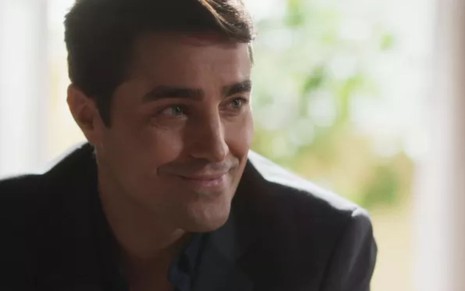 Danilo (Ricardo Pereira) está sentado e sorri em cena de Cara e Coragem, novela das sete da Globo