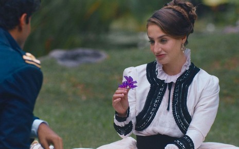 Leopoldina (Bruna Griphao) está sentada em jardim e segura uma flor em cena de Nos Tempos do Imperador