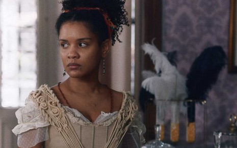 A atriz Heslaine Vieira, com expressão de raiva, olha para o lado esquerdo como a Zayla em um ateliê em cena de Nos Tempos do Imperador