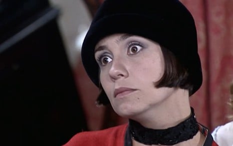 Adriana Esteves usa um vestido vermelho e um chapéu preto; ela tem os olhos arregalados e a boca apertada em cena de O Cravo e a Rosa