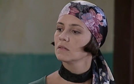 Adriana Esteves, caracterizada como Catarina, usa um vestido preto e um lenço florido; ela tem o semblante desconfiado em cena de O Cravo e a Rosa
