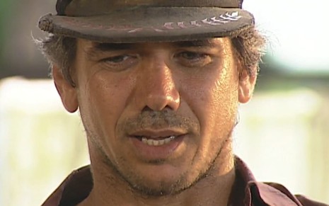 Jackson Antunes caracterizado como Regino: ator veste uma camiseta surrada e um boné. Ele tem o semblante abalado em cena de O Rei do Gado