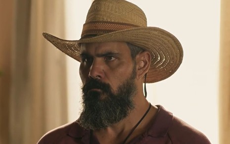 Juliano Cazarré, caracterizado como Alcides em Pantanal: ele tem a barba longa e grisalha. O ator veste um chapéu de palha e uma camisa polo vermelha; o semblante está furioso em cena de Pantanal