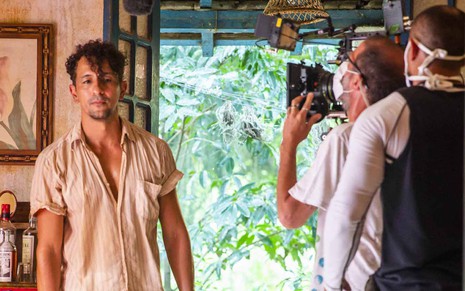 O ator Irandhir Santos exibe cabelo cacheado e camisa semiaberta ao ser acompanhado por técnicos em gravação de cena de Pantanal