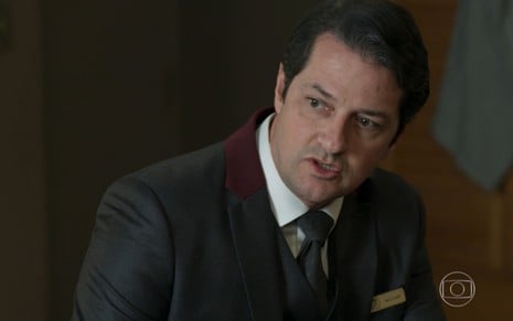 Marcelo Serrado interpretando Malagueta, vilão de Pega Pega. ele usa um terno preto com gravata azul e blusa branca e olha irritado ao lado direito.