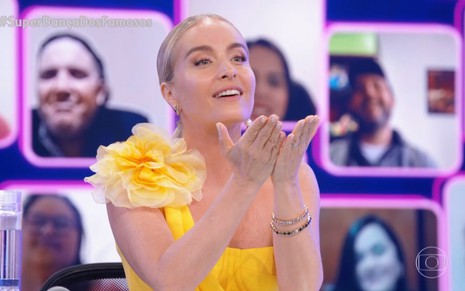 A apresentadora Angélica, com vestido amarelo de um ombro, faz um gesto simpático na bancada do júri da Super Dança dos Famosos