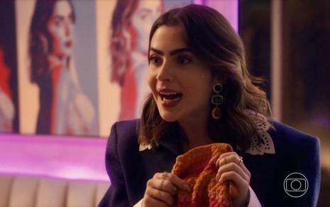 A atriz Jade Picon está segurando uma peça de crochê e faz uma cara de supresa com a boca aberta em cena da novela Travessia