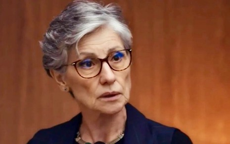 A atriz Cassia Kis usa óculos de grau e blazer azul marinho em cena de Travessia caracterizada como a personagem Cidália
