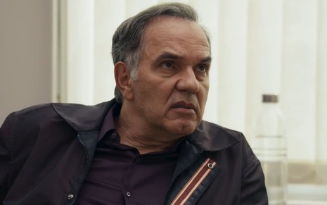 Humberto Martins caracterizado como Guerra em Travessia; ele exprime choque e usa uma camisa azul e uma jaqueta marrom em cena da novela
