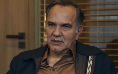 Humberto Martins caracterizado como Guerra em Travessia; ele tem o semblante preocupado e usa uma camiseta preta e uma jaqueta bege em cena da novela
