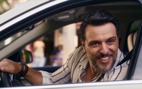 Rodrigo Lombardi está com uma blusa listrada ao volante de um automóvel em cena da novela Travessia