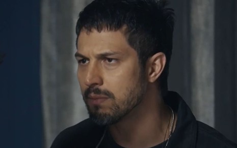 Romulo Estrela tem a expressão séria em cena de Travessia; ele usa uma camiseta preta, justa, e um colar de prata