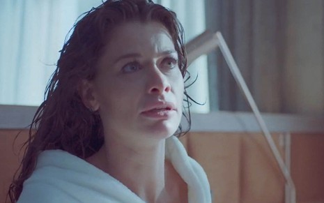 A atriz Alinne Moraes, de cabelos molhados, com um roupão branca, sentada na cama como a Bárbara de Um Lugar ao Sol