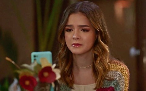 A atriz Mel Maia olha para um celular com cara de tristeza em cena da novela Vai na Fé como a personagem Guiga