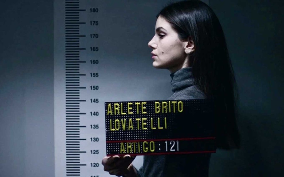 A atriz Camila Queiroz, como Angel, de perfil, tira uma fotografia ao ser fichada na prisão com uma placa em que se lê 'Arlete Pinheiro Lovatelli' em cena de Verdades Secretas 2