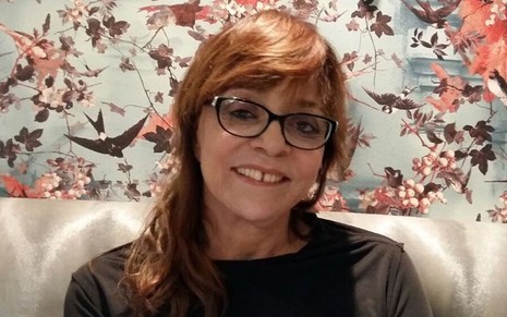 A autora de novelas Gloria Perez; ela está de óculos, sentada, posando para a foto e sorrindo