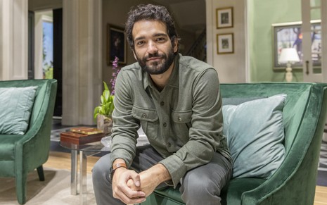 Retrato de Humberto Carrão, sorrindo, sentado em um sofá no cenário de Todas as Flores