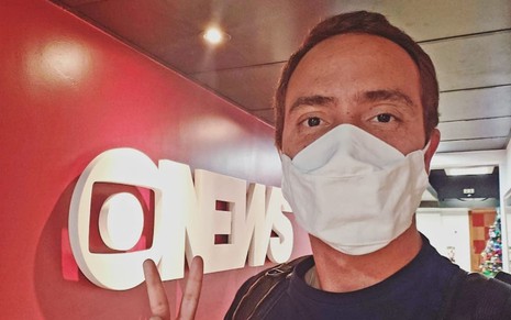 O repórter André Coelho posa em frente ao logo da GloboNews em foto publicada no Instagram; ele aparece de máscara
