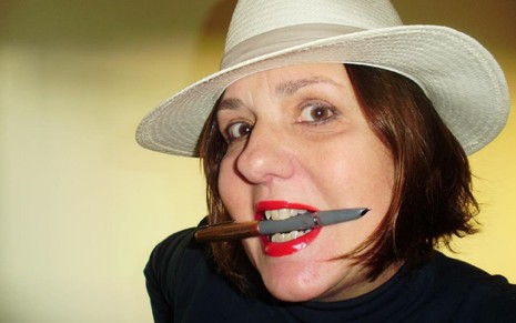 Jackie Vellego está de chapéu bege, ela segura uma caneta com os dentes e usa batom vermelho