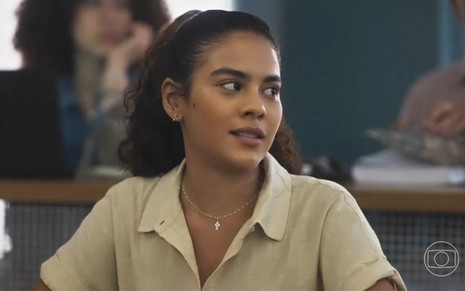 Em cena de Vai na Fé, Bella Campos está usando uma blusa bege e está olhando para alguém que está ao seu lado