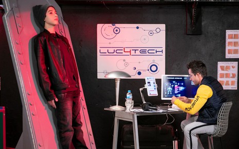 LUC2 (João Pedro Delfino) está numa maca elevada, enquanto Nicholas (Fábio Beltrão) mexe num computador