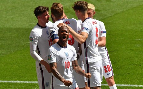 Raheem Sterling com uniforme todo branco da Inglaterra com os punhos cerrados e dando grito em celebração com jogadores da Inglaterra ao fundo se abraçando