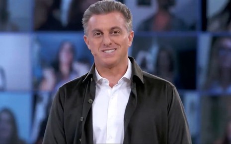 Luciano Huck veste um casaco preto com uma blusa marrom e sorri, convidando o público a assistir o seu primeiro programa nos domingos da Globo