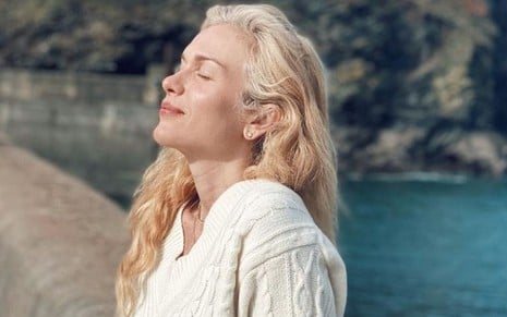 A atriz Ludmila Dayer de perfil, de olhos fechados, com blusa branca de tricô, em mureta de frente para uma praia