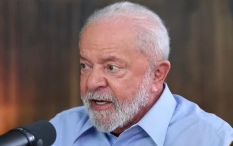 Luiz Inácio Lula da Silva usa camisa azul e está com expressão séria falando num microfone estilo podcast