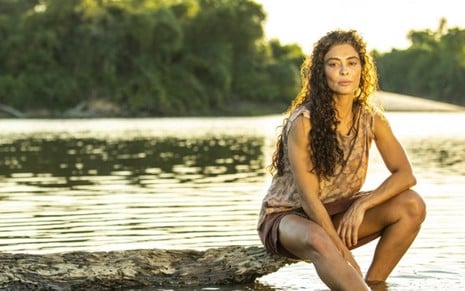 A atriz Juliana Paes como Maria Marruá em Pantanal; ela está sentada em um toco de árvore à beira de um lago