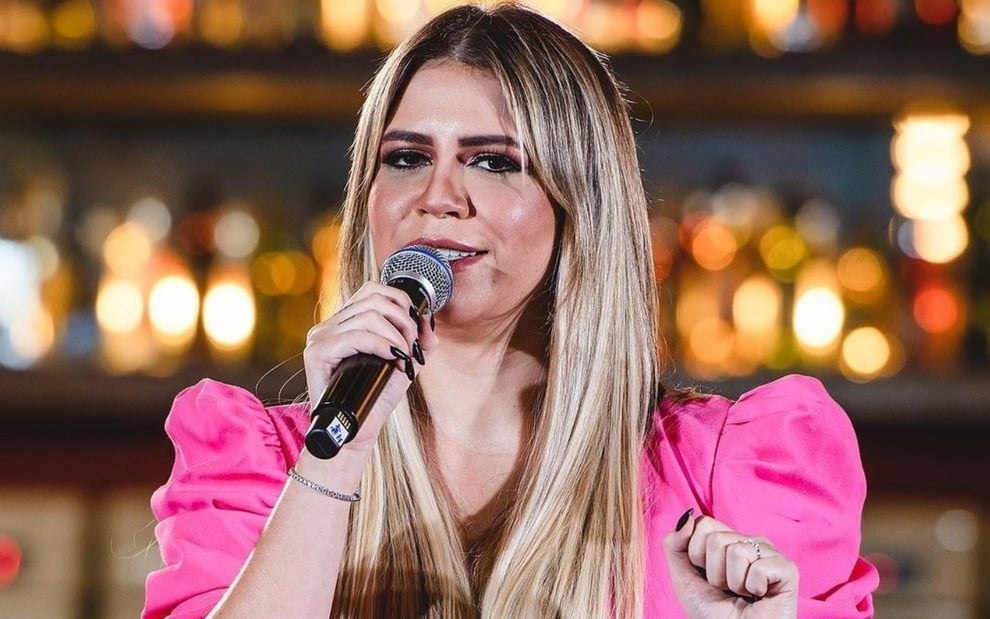 Marília Mendonça está com um microfone na mãe, vestida com uma camisa rosa, e canta em show