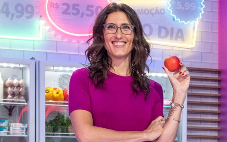 Paola Carosella em foto de divulgação do programa Minha Mãe Cozinha Melhor Que a Sua, da Globo, segurando uma maçã e sorrindo