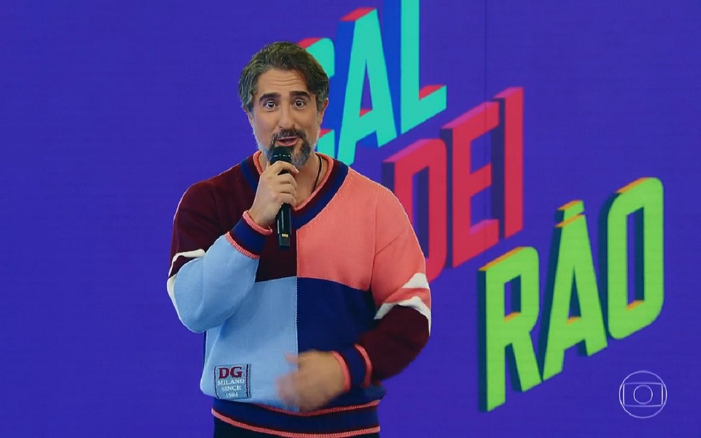 Marcos Mion usa uma camisa quadriculada, sorri e fala com o público do Caldeirão em sua estreia na Globo