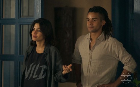 A atriz Julia Dalavia e o ator Lucas Leto como Guta e Marcelo em Pantanal; eles estão em pé, olhando para a frente com um leve riso no rosto, enquanto ela aponta a mão para o lado e ele encosta a mão na cintura