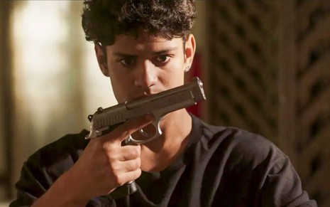 O ator Gabriel Santana como Renato em Pantanal; ele está segurando uma arma e olhando para ela com atenção