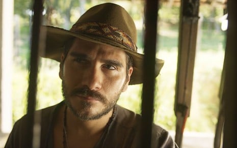 O ator Gabriel Sater como Trindade em Pantanal; ele está de chapéu, atrás de uma grande, olhando para cima com cara de desconfiado