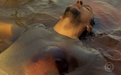 O ator Irandhir Santos como José Lucas em Pantanal; ele está caído no rio, de olhos fechados, com uma bala no peito