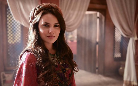 A atriz Thaís Melchior está com um vestido vermelho, longo, e um turbante no topo da cabeça que permite aos seus cabelos ficarem soltos como a Raquel em cena de Gênesis