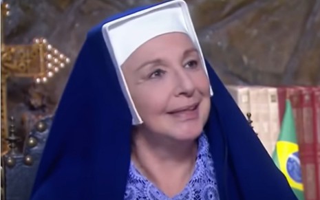 Eliana Guttman grava cena vestida de freira, sorrindo, como Madre Superiora em Carinha de Anjo, do SBT