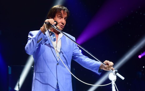 Em cim do palco, Roberto Carlos canta, segurando um microfone, vestindo um blazer e uma calça azuis
