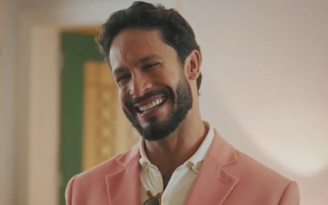 O ator Rainer Cadete usa blazer rosa e camisa branca em cena da novela Terra e Paixão na qual sorri como Luigi