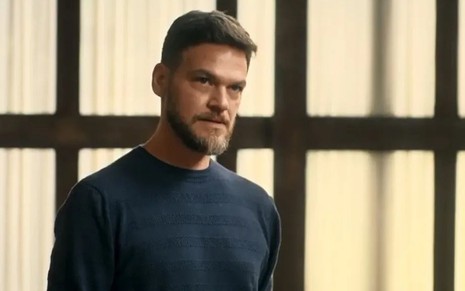 Em cena de Vai na Fé, Emilio Dantas usa uma blusa lsitrada azul e preta e olha para alguém com raiva