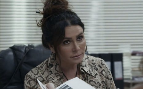 Com um coque no alto da cabeça, Giovanna Antonelli usa blusa estampada, está na delegacia, com sua persiana ao fundo, mostrando um documento a alguém na sua frente