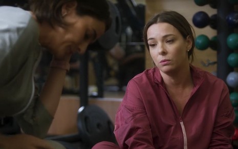 Em cena de Vai na Fé, Regiane Alves está com roupa esportiva rosa, sentada no chão da academia, ao lado de Priscila Sztejnman, que sorri