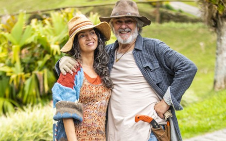 Claudia Ohana e Zécarlos Machado usam chapéu e posam no jardim de uma pousada, cenário de Vai na Fé