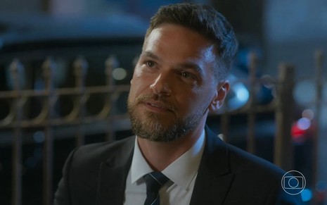 Em cena de Vai na Fé, Emilio Dantas está usando terno e gravata e está olhando para alguém