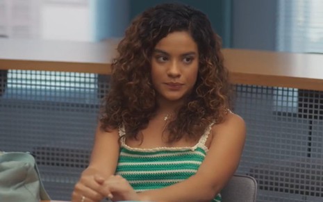 A atriz Bella Campos com expressão séria, blusa sem mangas verde e branca, em sala de aula em cena de Vai na Fé