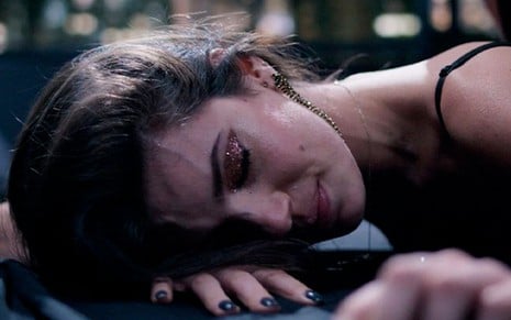 A atriz Camila Queiroz como Angel em Verdades Secretas 2; ela está deitada de bruços, com a cabeça em cima da mão e cara de cansada
