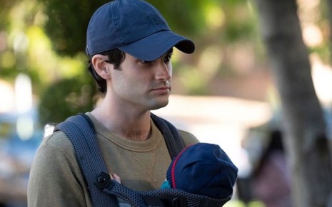 Joe (Penn Badgley) usa um boné e carrega um bebê em cena de You, série da Netflix